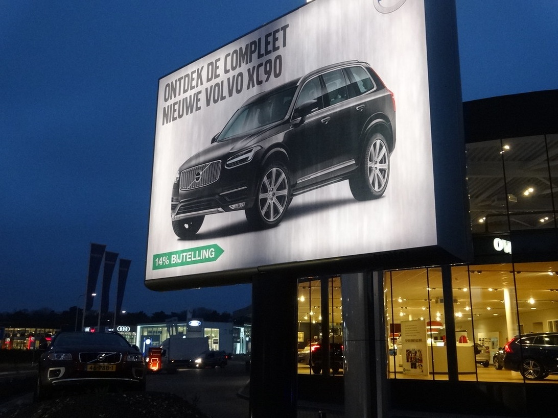 En stor reklamskylt för en Volvo XC90 lyser upp i skymningen utanför en modern bilhall.